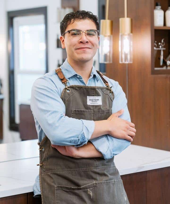 A barber named Daniel from Barber Haus in Naperville standing confidently with arms crossed, wearing glasses and a blue shirt under a brown apron with the 'Victory' brand logo on it. He is in a modern barbershop with stylish lighting and wooden decor.