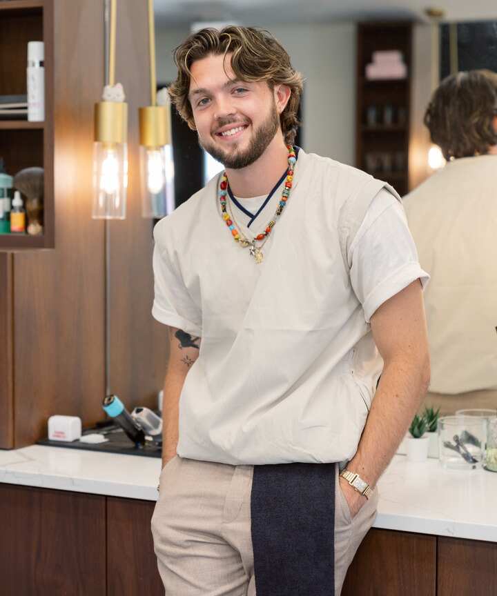 Danny the Barber in Naperville wearing a beige shirt with colorful beaded necklaces, stands casually in a modern barber shop with his hands in his pockets.