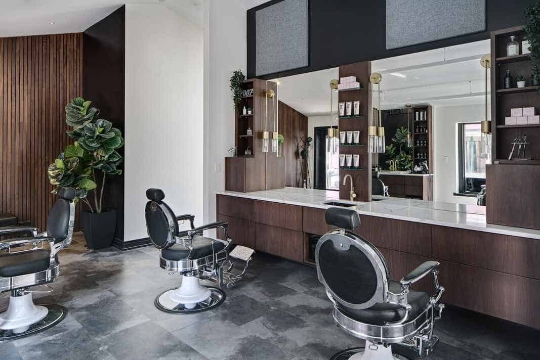 Interior of Barber Haus in Naperville, featuring modern barber chairs with chrome and black leather finishes. The sleek, contemporary design includes dark wood paneling, large mirrors, and minimalistic decor with green plants and stylish shelving for grooming products. The space is well-lit, with pendant lights adding a warm ambiance to the clean, polished atmosphere.