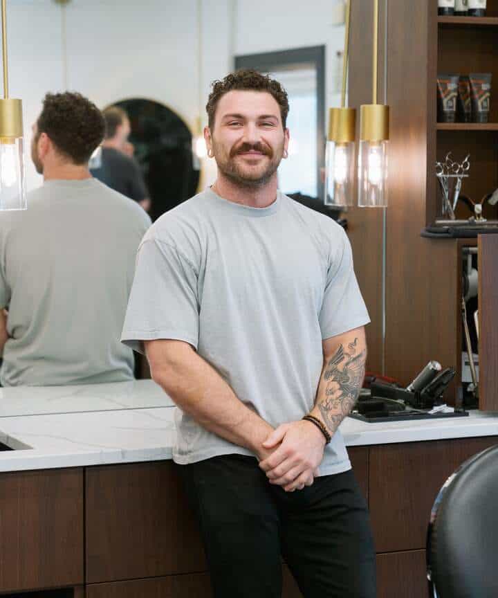 Jonathan a Barber in Naperville wearing a light gray t-shirt, smiles while standing with his hands clasped in front of him in a modern barber shop.